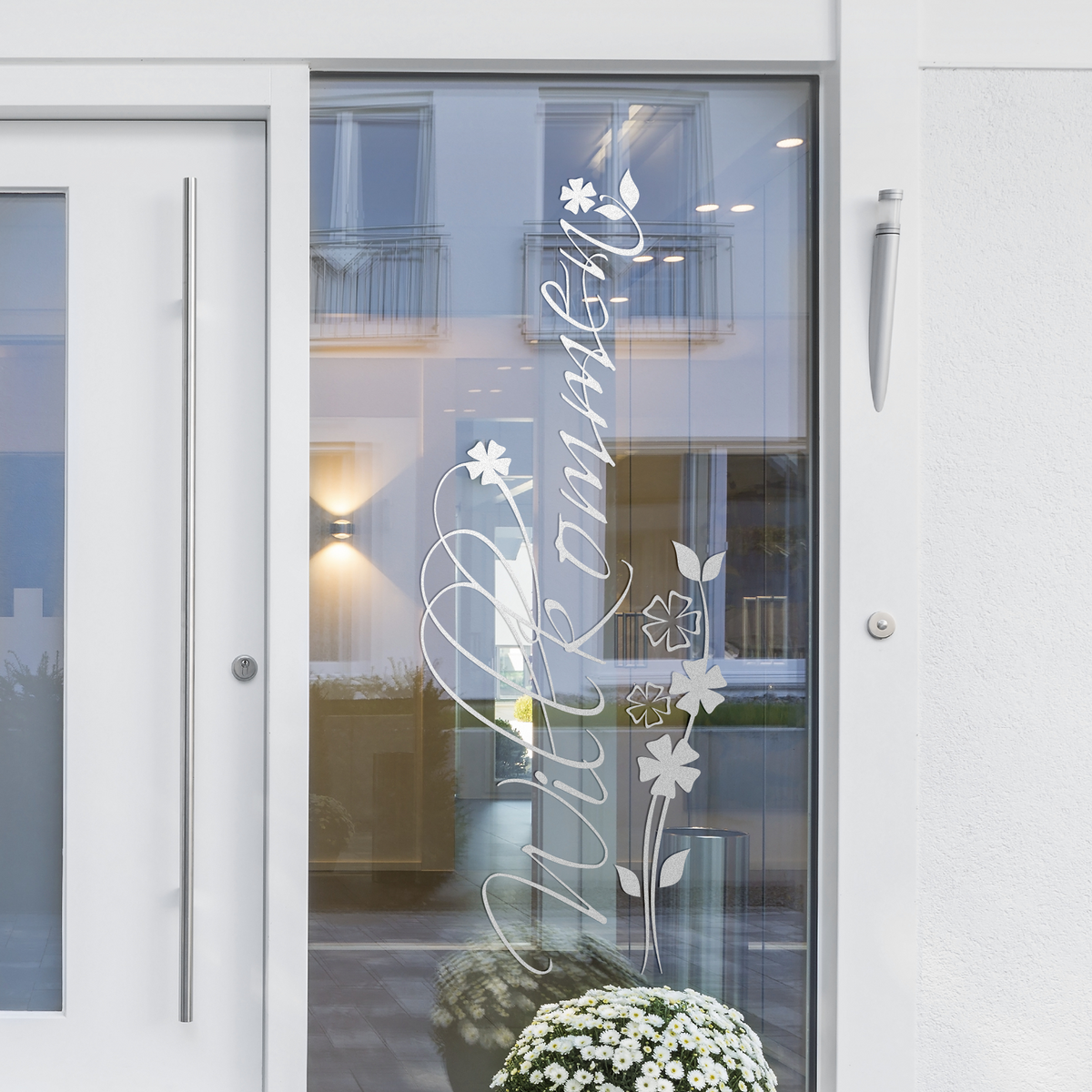 Willkommen-Schriftzug in Weiß auf Glastür mit Blumenmotiven.