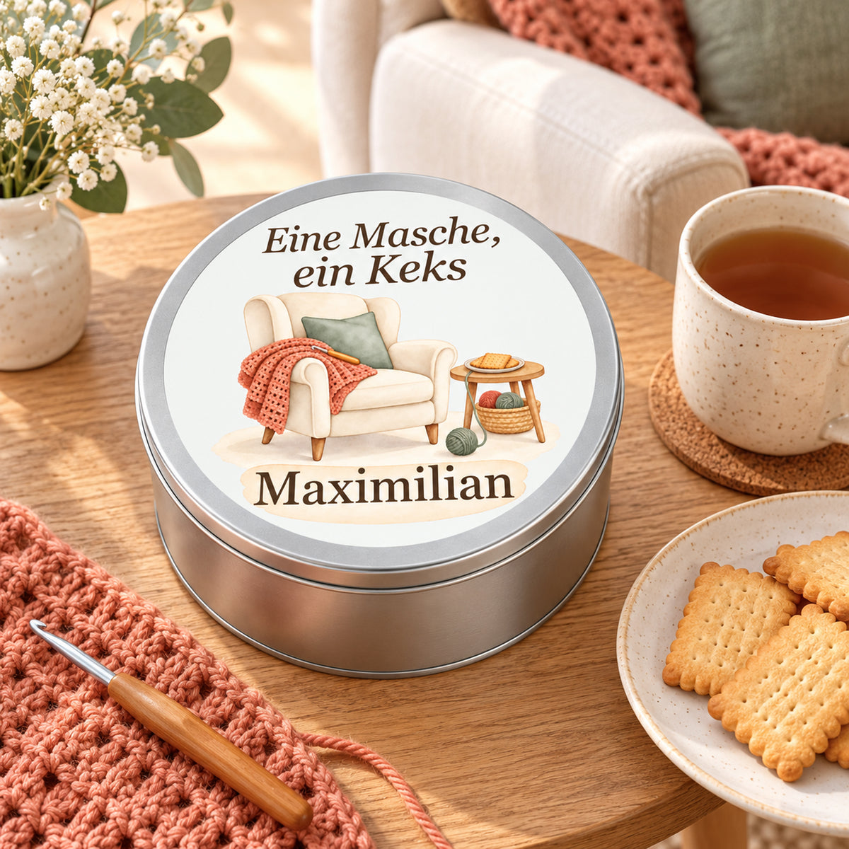 Geschlossene Keksdose mit Häkelmotiv und Namensfeld neben Tasse, Keksen und Häkelmaterial.