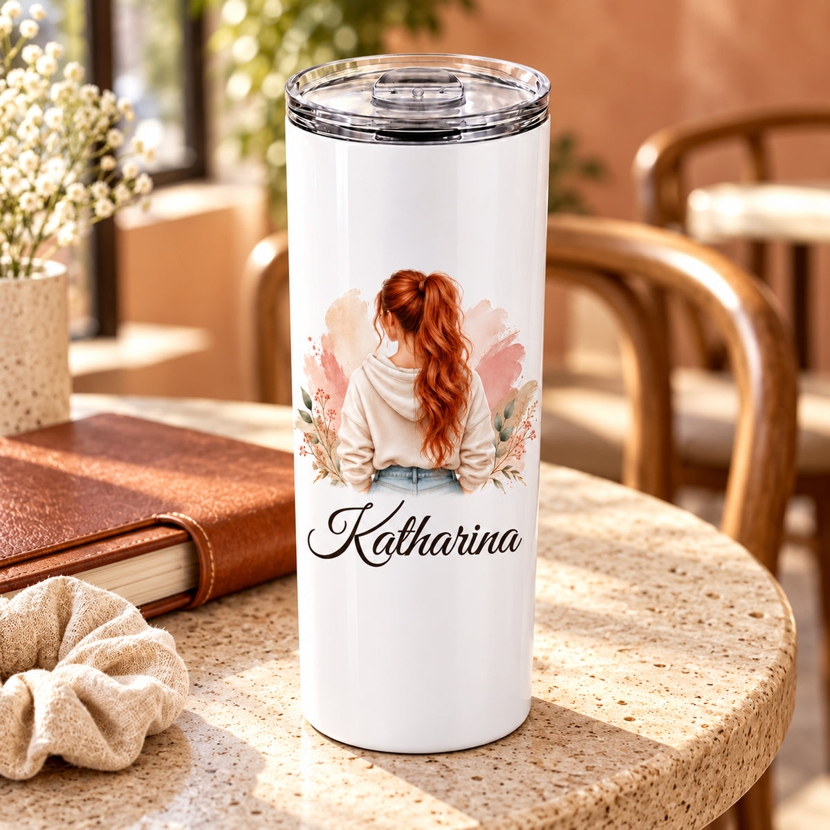 Thermobecher mit Namen und Frauenmotiv mit kupferrotem Pferdeschwanz und cremefarbenem Hoodie in einer Cafészene.