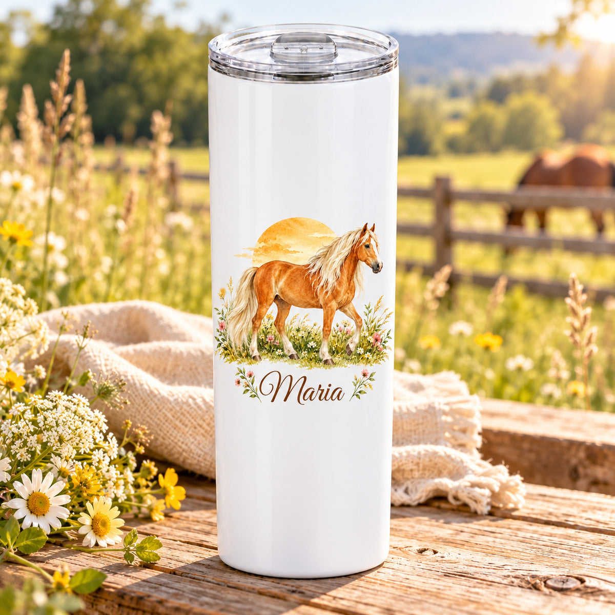 Weißer Thermobecher mit einem Haflinger vor einem Sonnenbogen und Wunschname auf einem Holztisch vor einer Pferdekoppel.