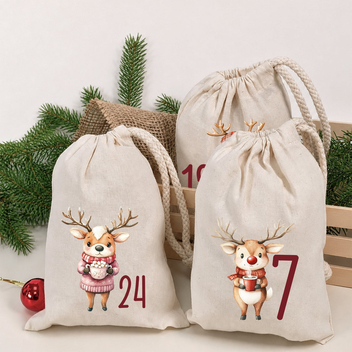 Drei naturfarbene Adventskalender-Stoffbeutel mit Rentiermotiven und den Zahlen 24, 18 und 7 vor Tannenzweigen.