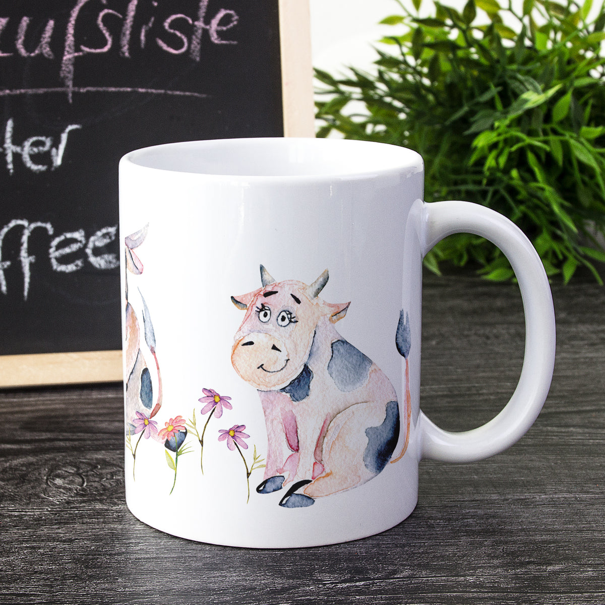 Weißer Kaffeebecher mit bunten Kuh-Motiven und Blumen.