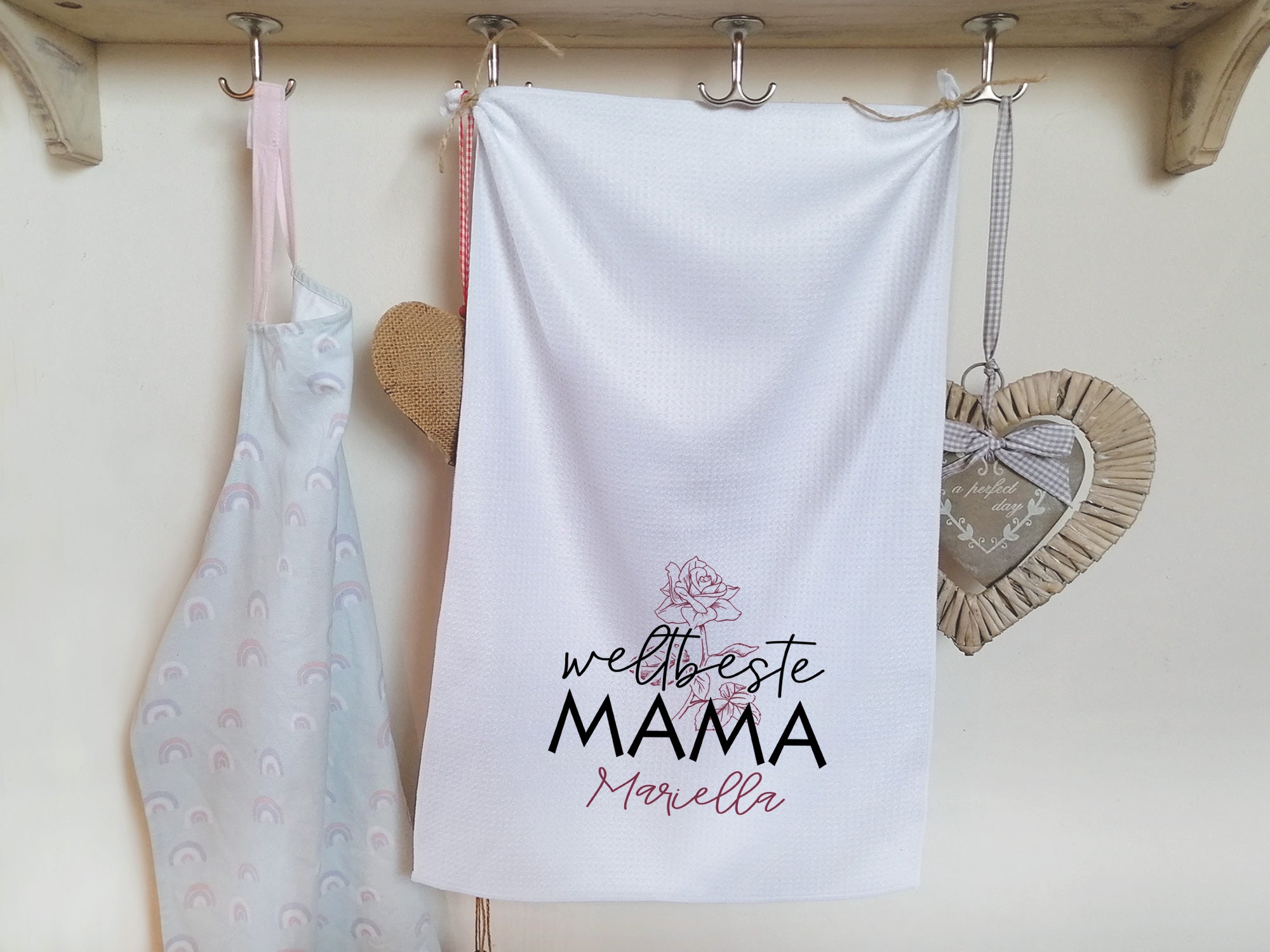 Weißes Geschirrtuch mit schwarzem Text "weltbeste MAMA" und rosa Blume.