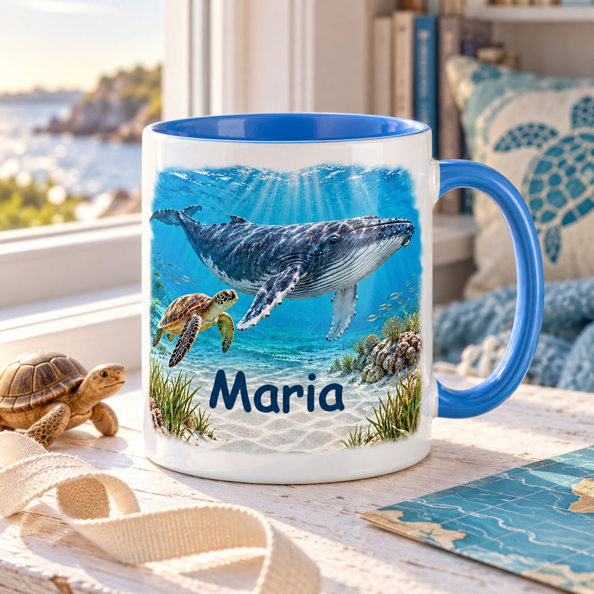 Wal-Tasse mit Meeresschildkröte und Name Maria auf einer Fensterbank mit Meerblick, Büchern und Holzschildkröte