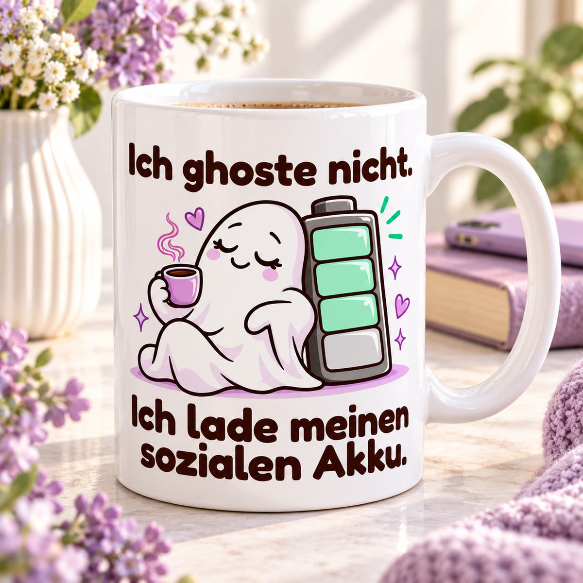 Weiße Keramiktasse mit dem Spruch „Ich ghoste nicht. Ich lade meinen sozialen Akku.“ und farbigem Cartoonmotiv in einer Leseecke mit Buch.