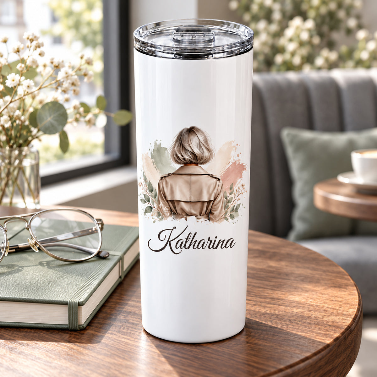 Thermobecher mit Namen und Frauenmotiv mit silbergrauem Bob und taupefarbenem Trenchcoat in einer Cafészene.