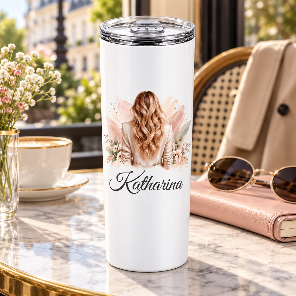 Thermobecher mit Namen und Frauenmotiv mit blonden Wellen und beigem Blazer in einer Cafészene.