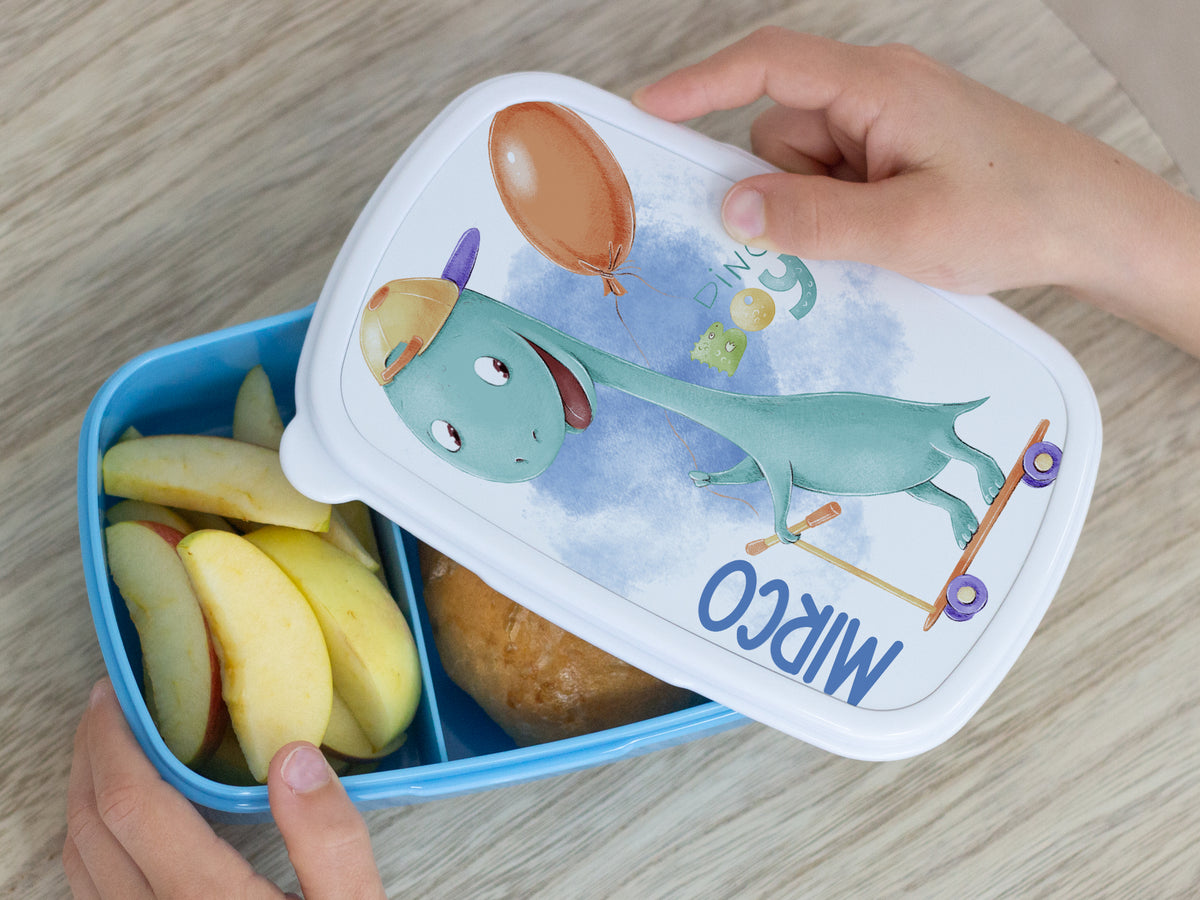 Kinderteller mit Dinosaurier-Motiv, blaue Box, Äpfel und Brötchen.