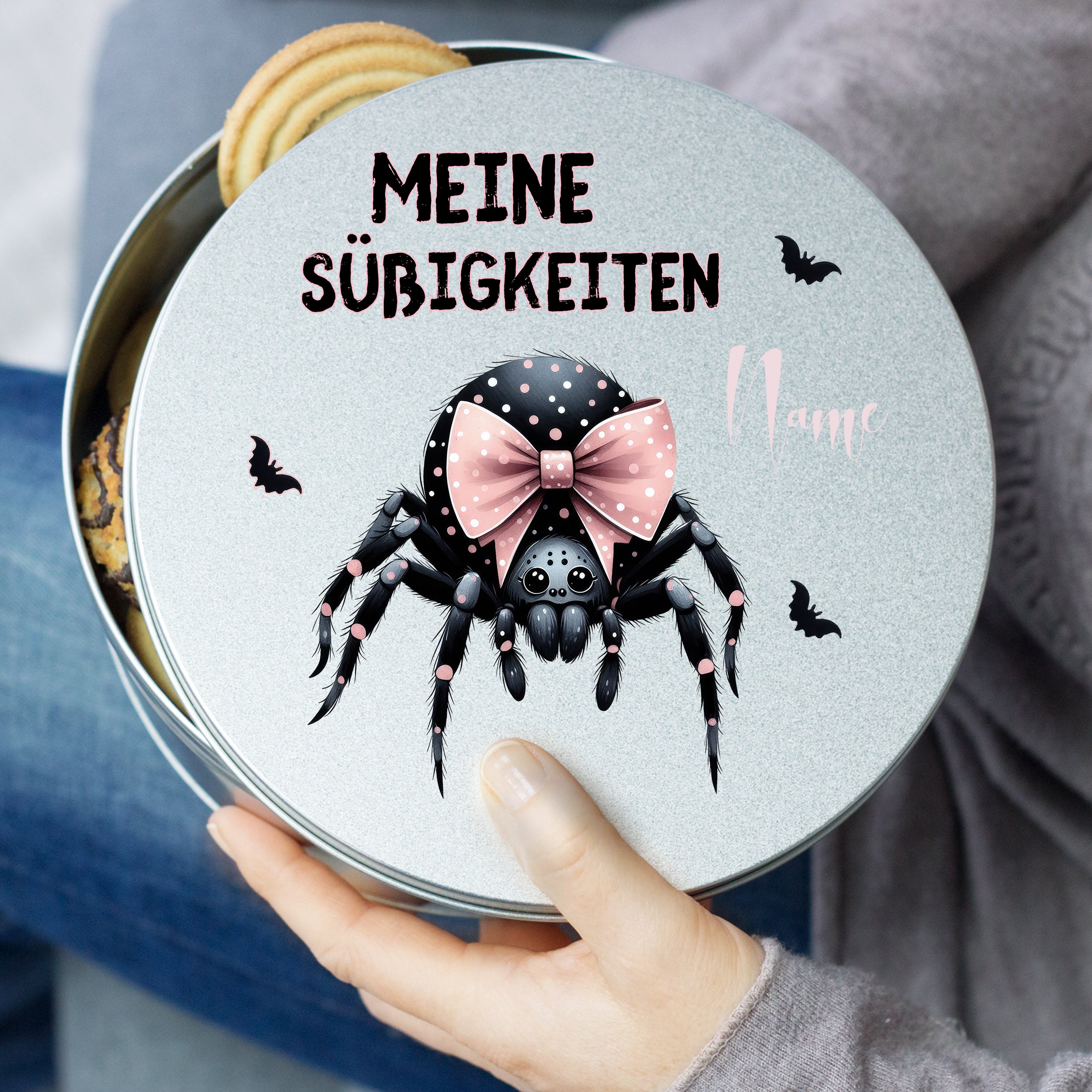 Graue Keksdose mit schwarzer Spinne und pinkem Schleifenmotiv.