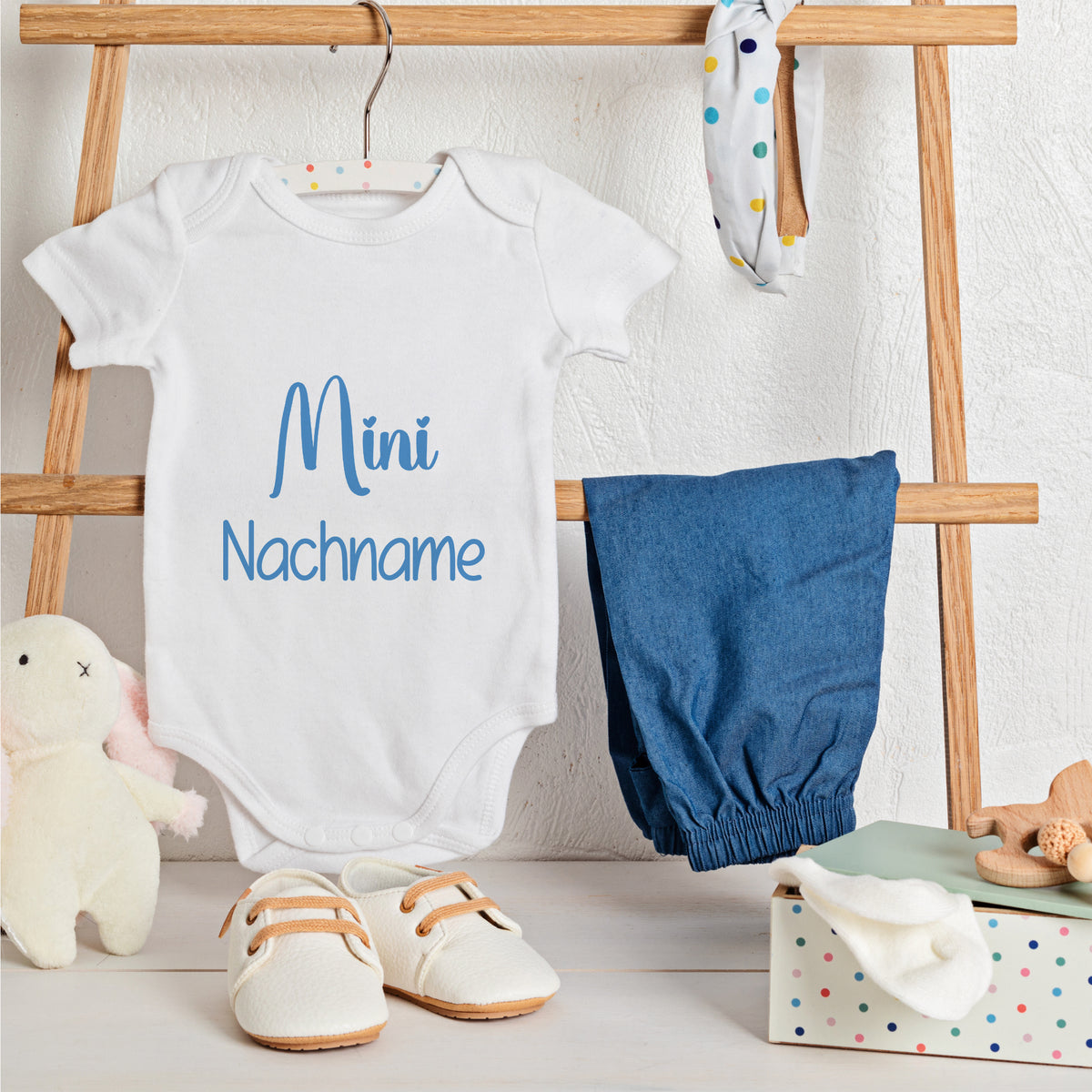 Weißer Babybody mit blauer Schrift "Mini Nachname", blaue Hose.