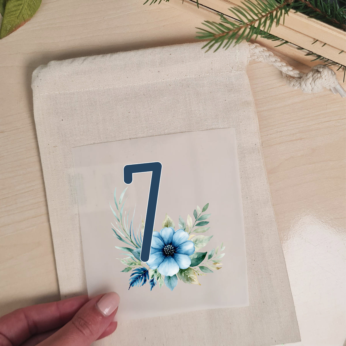 Blaues Blumenmotiv mit Zahl 7 wird auf einen naturfarbenen Stoffbeutel aufgelegt