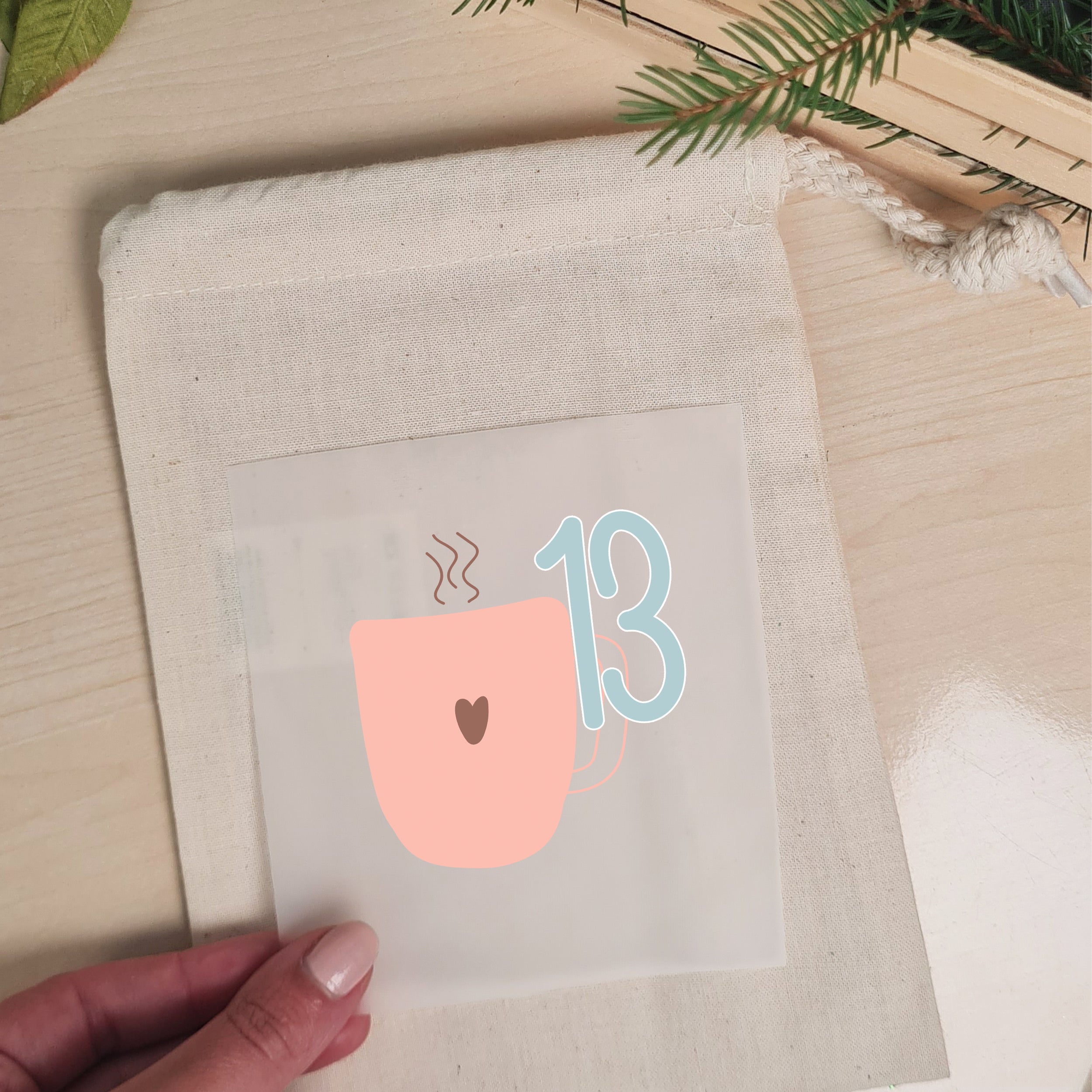 Pastellfarbenes Weihnachtsmotiv mit Zahl 13 wird auf einen naturfarbenen Stoffbeutel aufgelegt