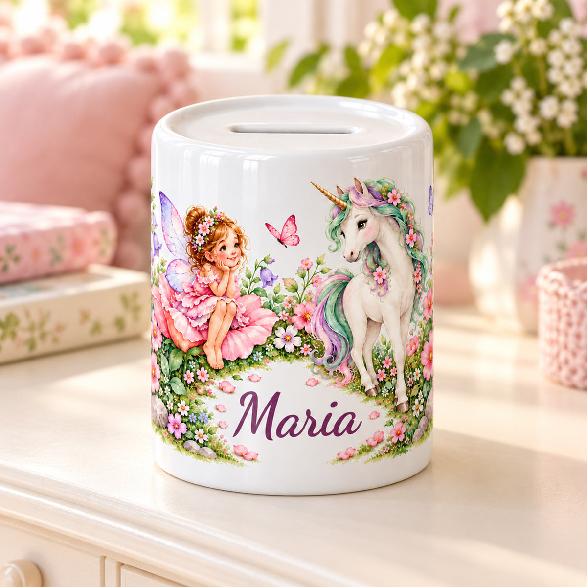 Personalisierte Feen-Einhorn-Spardose auf einer hellen Kommode neben rosa Kissen und Blumen.