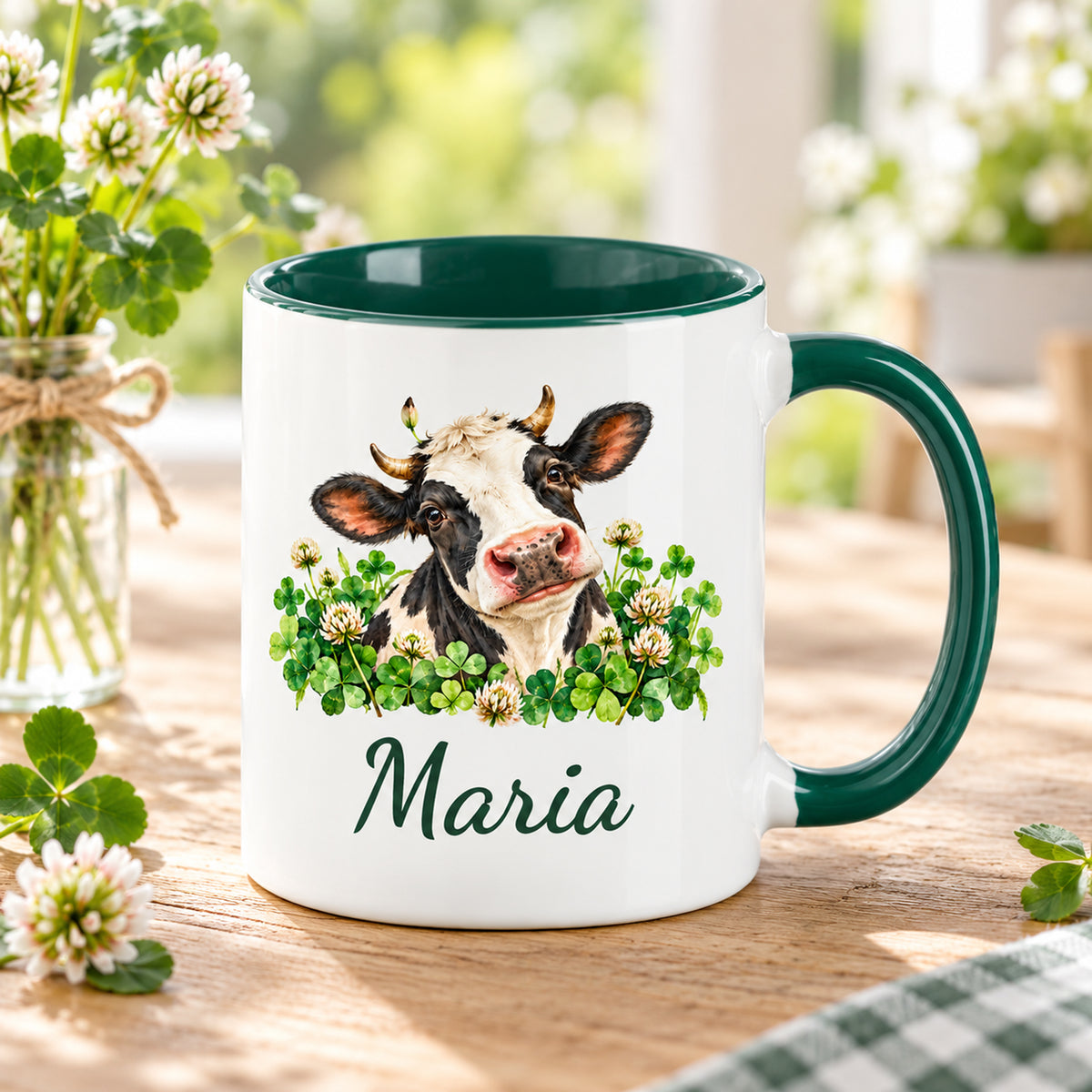 Kuh-Tasse mit schwarz-weißer Kuh zwischen Kleeblättern und Blüten und Wunschname auf einem Holztisch mit Wiesenblumen