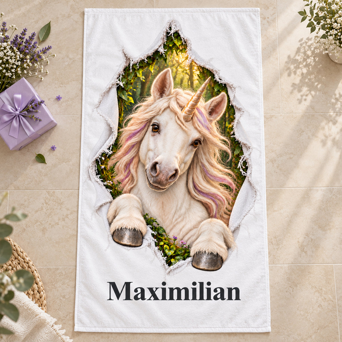 Weißes personalisiertes Handtuch mit Einhorn-Motiv im Stoffriss-Design auf hellem Steinboden neben einer lilafarbenen Geschenkbox und Blumen.